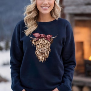May include: Navy blue sweatshirt featuring a Highland cow graphic with a pink bow. The cow has long, shaggy brown fur and horns. The sweatshirt is paired with blue jeans and a white knit beanie. The background shows a snowy landscape and a fireplace.
