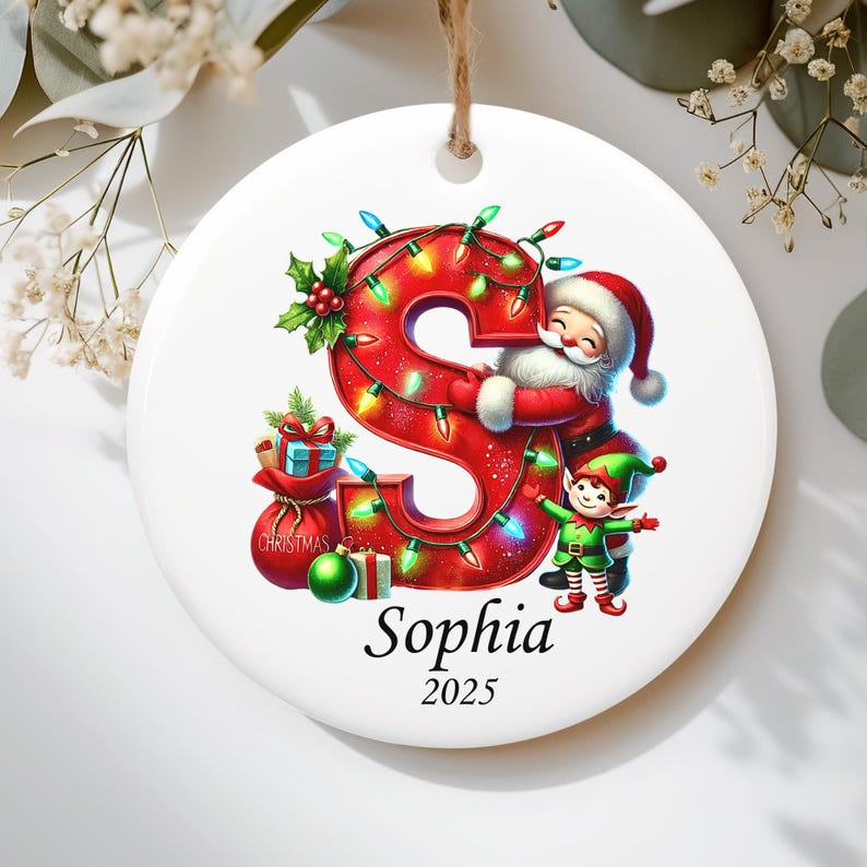 May include: A round white ceramic Christmas ornament featuring a large red letter S wrapped in colorful string lights. A Santa Claus and an elf stand beside the letter, with a red gift sack and ornaments below. The name Sophia and the year 2025 are printed.