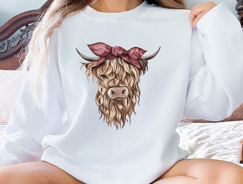 May include: White sweatshirt featuring a watercolor illustration of a Highland cow with a pink bandana. The cow has long, shaggy brown fur, dark horns, and a light brown face. The sweatshirt is a crew neck style.
