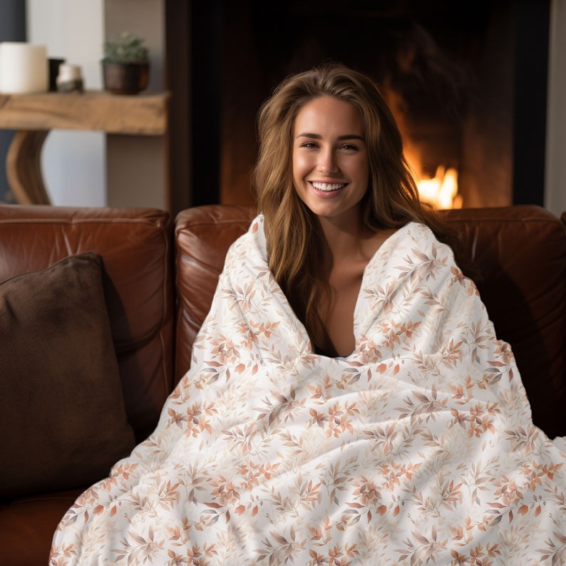 May include: A woman sitting on a brown leather couch, wrapped in a white throw blanket with a brown floral pattern. The blanket has a soft, cozy feel and is perfect for snuggling up on a cold day.