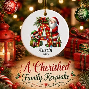 May include: A round white ceramic Christmas ornament featuring a large red letter A decorated with colorful string lights, a Santa Claus figure, and an elf. The text Austin and 2025 are printed below the letter. It hangs by a gold string.