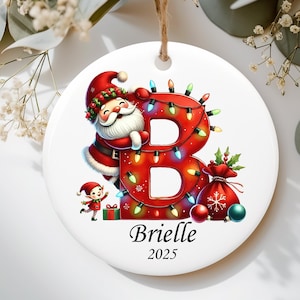 May include: A round white ceramic ornament featuring a large red letter B wrapped in colorful Christmas lights. A smiling Santa Claus leans against the letter, accompanied by a small elf, a gift box, and a red sack. The name Brielle and year 2025 are printed below.