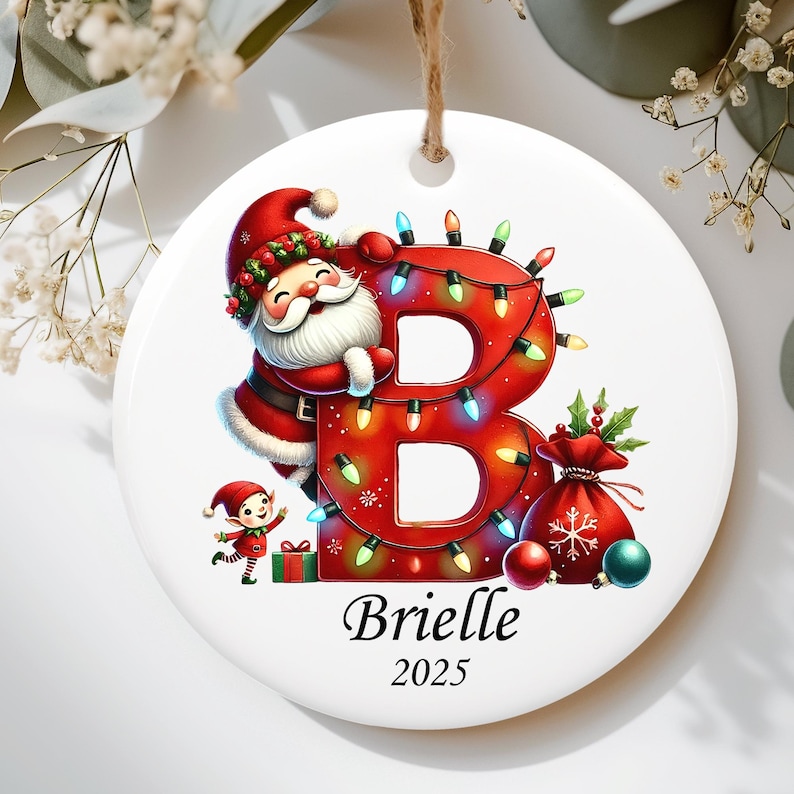 May include: A round white ceramic ornament featuring a large red letter B wrapped in colorful Christmas lights. A smiling Santa Claus leans against the letter, accompanied by a small elf, a gift box, and a red sack. The name Brielle and year 2025 are printed below.