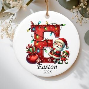 May include: A round white ceramic Christmas ornament featuring a large red letter E wrapped in colorful string lights. Beside the letter stands Santa Claus and a small elf next to a red gift sack. The name Easton and the year 2025 are printed below.