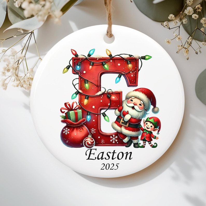 May include: A round white ceramic Christmas ornament featuring a large red letter E wrapped in colorful string lights. Beside the letter stands Santa Claus and a small elf next to a red gift sack. The name Easton and the year 2025 are printed below.