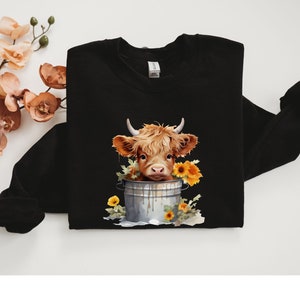 May include: Black sweatshirt with a graphic of a brown and white Highland cow with a yellow flower crown in a metal bucket.