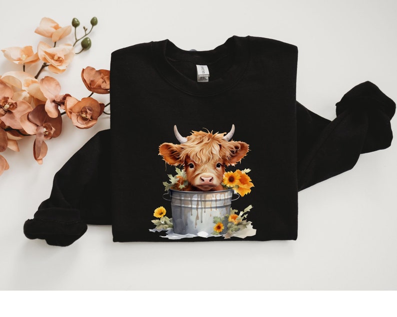 May include: Black sweatshirt with a graphic of a brown and white Highland cow with a yellow flower crown in a metal bucket.
