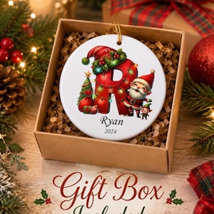May include: A round white ceramic Christmas ornament featuring a large red letter R decorated with green holly, a small Christmas tree, and a cheerful Santa Claus figure. The name Ryan and the year 2024 are printed in black below the letter R.
