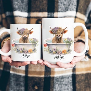 May include: Two white ceramic mugs with a Highland cow in a bathtub surrounded by flowers. The mugs are personalized with the name "Ashlyn".
