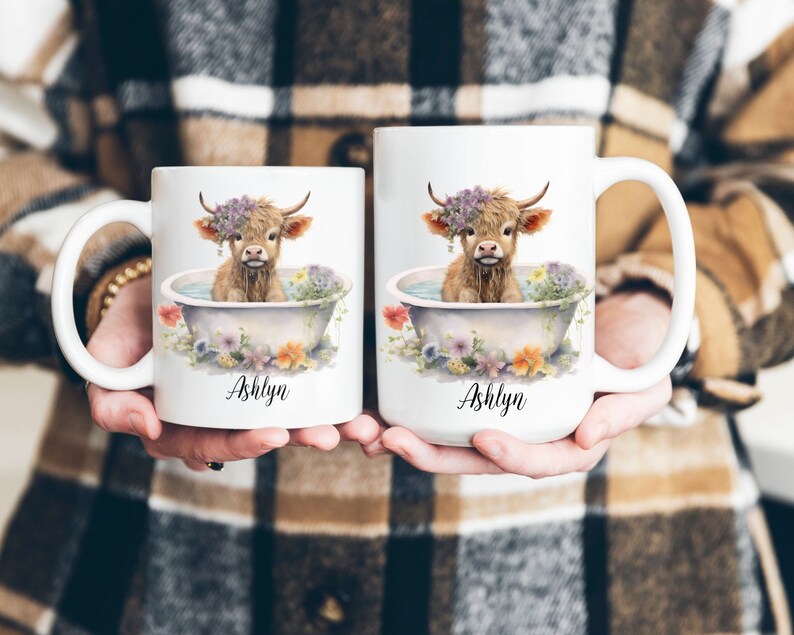 May include: Two white ceramic mugs with a Highland cow in a bathtub surrounded by flowers. The mugs are personalized with the name "Ashlyn".