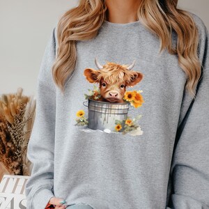 May include: A gray sweatshirt with a graphic of a brown and white Highland cow in a metal bucket with yellow sunflowers.