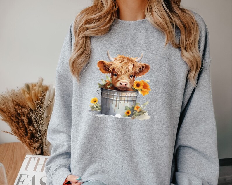 May include: A gray sweatshirt with a graphic of a brown and white Highland cow in a metal bucket with yellow sunflowers.