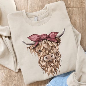 May include: A beige sweatshirt featuring a watercolor illustration of a Highland cow wearing a pink bandana. The cow has long, shaggy brown fur, dark horns, and a detailed face. The sweatshirt is folded on a wooden surface with a pair of blue jeans.