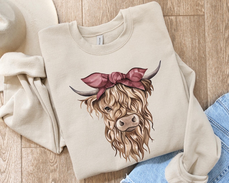 May include: A beige sweatshirt featuring a watercolor illustration of a Highland cow wearing a pink bandana. The cow has long, shaggy brown fur, dark horns, and a detailed face. The sweatshirt is folded on a wooden surface with a pair of blue jeans.