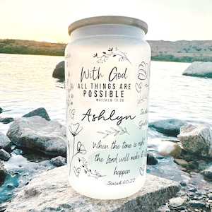 May include: A clear glass jar with a wooden lid, featuring the text "With God All Things Are Possible" and the name "Ashlyn". Floral and heart illustrations surround the text. The jar is sitting on a rock near water.