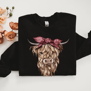 May include: Black sweatshirt featuring a detailed illustration of a Highland cow wearing a pink bandana. The cow has long, shaggy, light brown fur, a brown nose, and curved horns. The design is centered on the front of the long-sleeved garment.