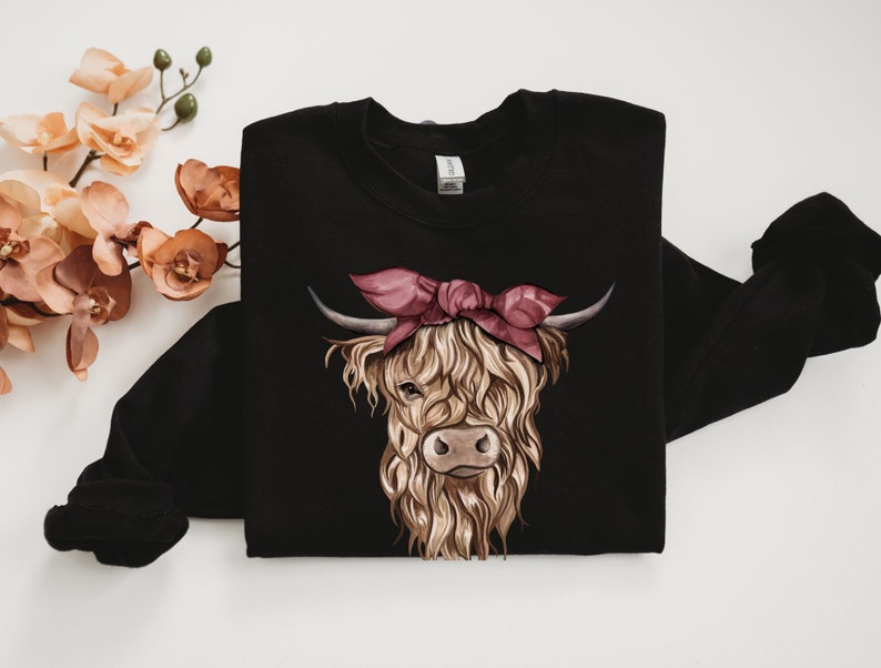 May include: Black sweatshirt featuring a detailed illustration of a Highland cow wearing a pink bandana. The cow has long, shaggy, light brown fur, a brown nose, and curved horns. The design is centered on the front of the long-sleeved garment.