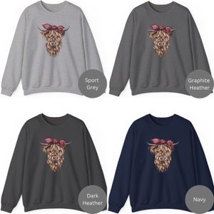 May include: Four sweatshirts in sport grey, graphite heather, dark heather, and navy. Each features a detailed illustration of a Highland cow with a pink bandana. The cow's fur is a mix of brown and tan, with a pink bandana.