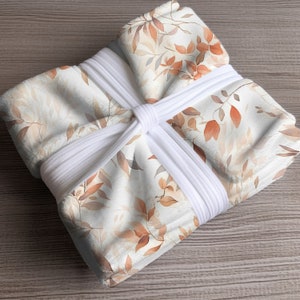 May include: A white throw blanket with a delicate watercolor pattern of brown and orange leaves. The blanket is tied with white ribbon.