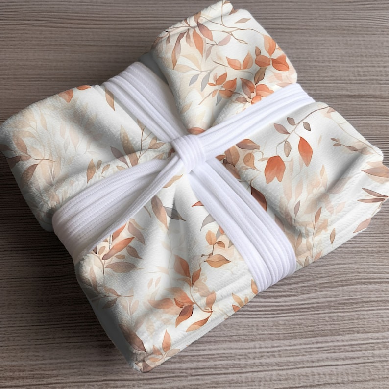 May include: A white throw blanket with a delicate watercolor pattern of brown and orange leaves. The blanket is tied with white ribbon.