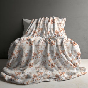 May include: A white throw blanket with a brown and orange floral pattern. The blanket is folded and draped over a white pillow.