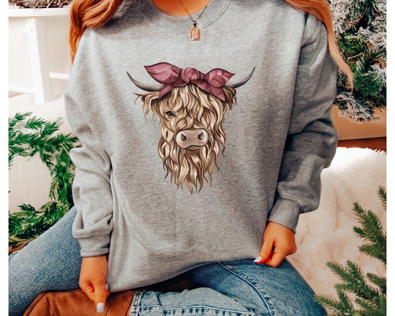 May include: A heather gray sweatshirt featuring a detailed illustration of a Highland cow. The cow has long, shaggy fur, horns, and a pink bow. The sweatshirt is a comfortable, casual style.