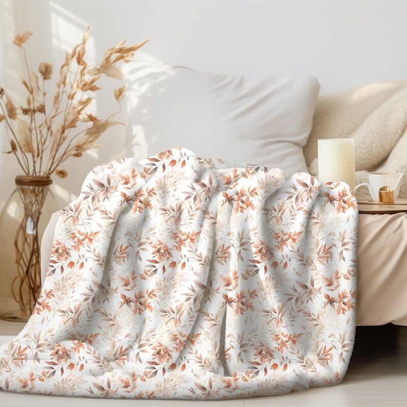 May include: A white throw blanket with a watercolor floral pattern featuring brown and orange leaves. The blanket is folded and draped over a white couch.