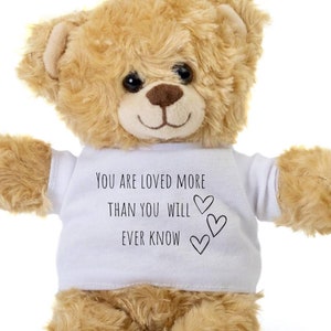Può includere: Un orsacchiotto marrone che indossa una maglietta bianca con il testo "You are loved more than you will ever know" e tre piccoli cuori.
