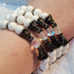 May include: Three beaded bracelets featuring smooth white spherical beads, translucent iridescent beads, and dark metallic dragon head charms. The bracelets are strung with small silver-toned spacer beads between the white and dragon components.
