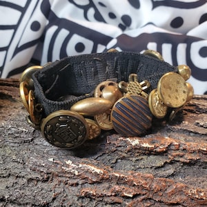 May include: A black fabric bracelet adorned with various antique brass-colored buttons and charms. The bracelet features a mix of round, oval, and uniquely shaped embellishments, creating a vintage aesthetic. The background includes a black and white patterned fabric.