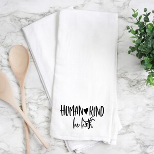 May include: White kitchen towel with black text that reads "HUMAN♥KIND be both".