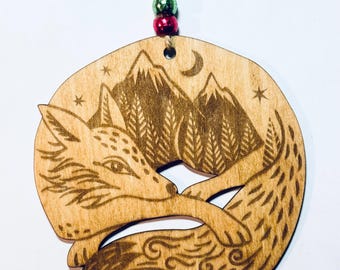 May include: Wooden ornament featuring a sleeping fox curled up in front of a mountain scene with a crescent moon and stars. The ornament is made of light-colored wood and has a natural finish. A jute string with green and red beads is attached for hanging.