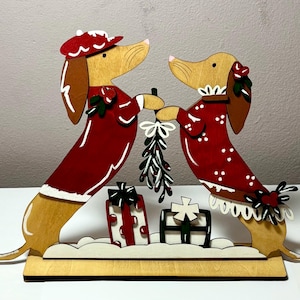 May include: Wooden Christmas decoration featuring two dachshund dogs in red outfits, holding mistletoe. The dogs stand on a wooden base with decorative presents and snow. The dogs have brown ears and noses, and wear hats and dresses.