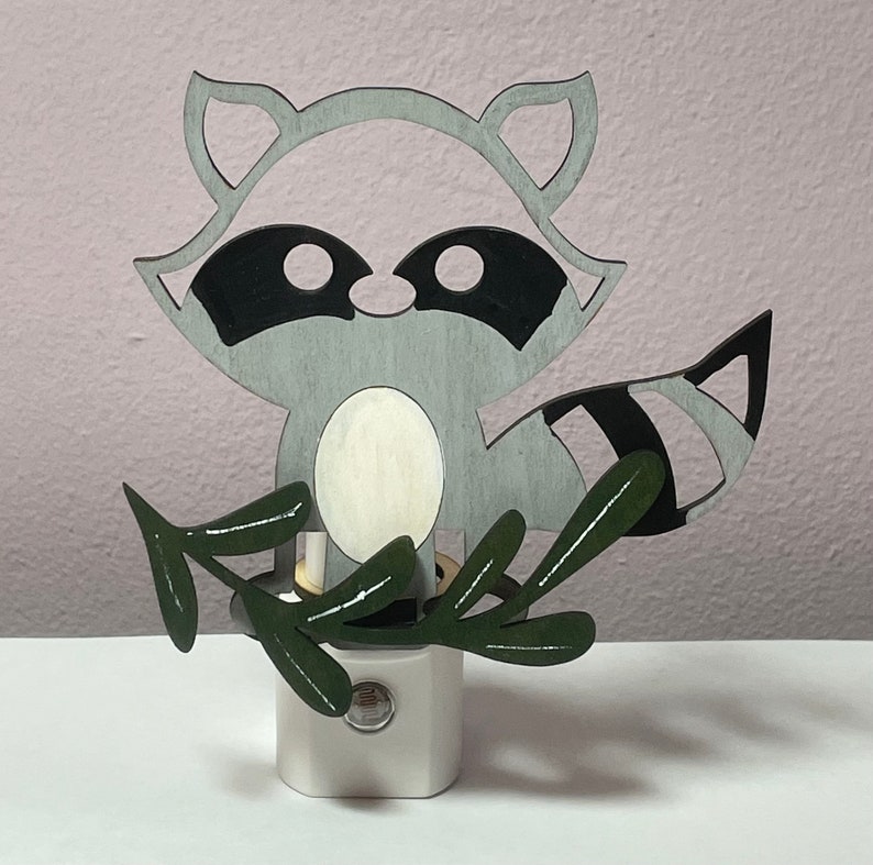 May include: A gray raccoon night light with a white belly and black eyes. The raccoon is holding a green leafy branch in its paws. The night light is on a white base.