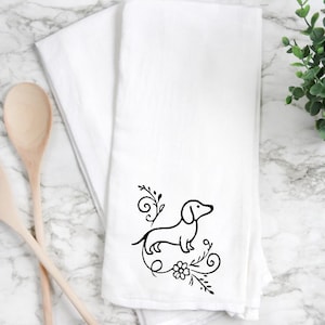 May include: White kitchen towel with a black line drawing of a dachshund dog surrounded by floral and swirl designs. The towel is folded and placed on a marble surface, with wooden spoons visible on the left side.