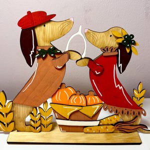 Puede incluir: Un recorte decorativo de madera con dos perros salchicha sobre una base. El perro de la izquierda lleva una boina roja y un su&eacute;ter marr&oacute;n, mientras que el de la derecha lleva una flor amarilla y un vestido rojo. Sostienen un hueso de la suerte blanco.