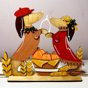 Puede incluir: Una decoraci&oacute;n de madera recortada que muestra dos perros salchicha frente a frente sosteniendo un hueso de la suerte blanco. El perro de la izquierda lleva una boina roja y un collar verde, el de la derecha una flor amarilla. Entre ellos hay una cesta de calabazas naranjas.