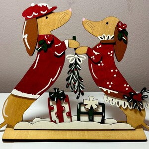 May include: Wooden Christmas decoration featuring two dachshund dogs in red outfits, holding mistletoe. The dogs stand on a wooden base with two gift boxes. The dogs have brown ears and noses, and wear hats and collars.