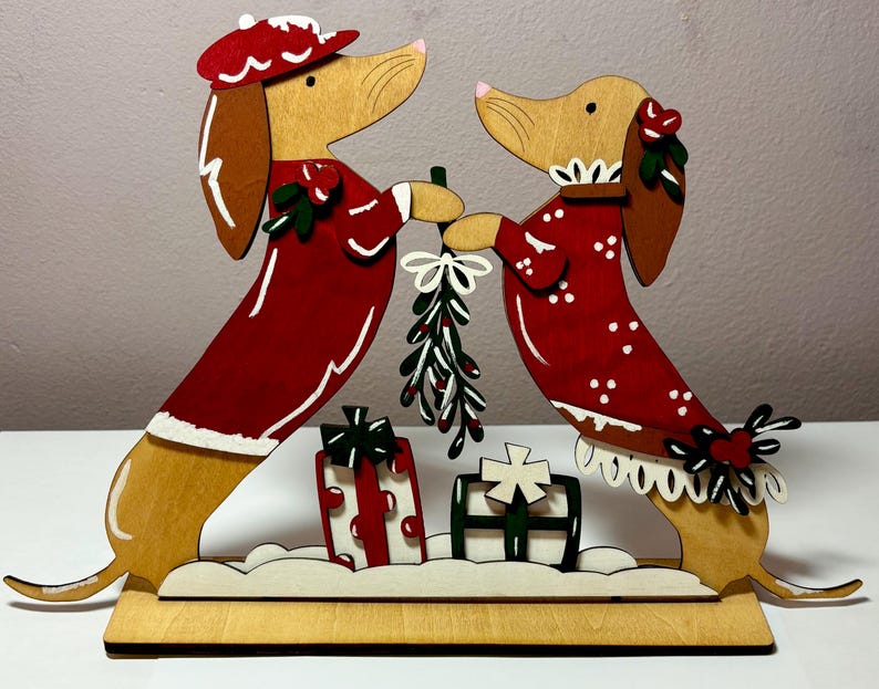 May include: Wooden Christmas decoration featuring two dachshund dogs in red outfits, holding mistletoe. The dogs stand on a wooden base with two gift boxes. The dogs have brown ears and noses, and wear hats and collars.