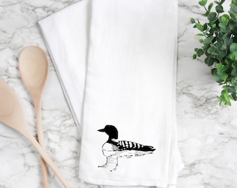May include: White kitchen towel with a black and white illustration of a loon. The loon is depicted in a simple, artistic style, with the bird floating on water. The towel is folded and placed on a marble surface.