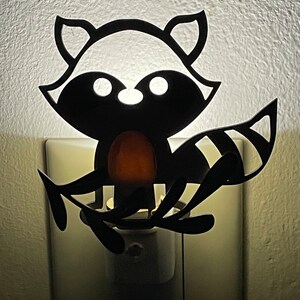 May include: A black silhouette of a raccoon with a light bulb in its belly, plugged into an outlet. The raccoon is holding a branch in its paws.
