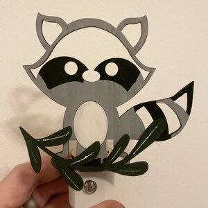 May include: A gray and black raccoon night light with a white belly and a green leaf design. The raccoon is holding a light bulb in its paws.
