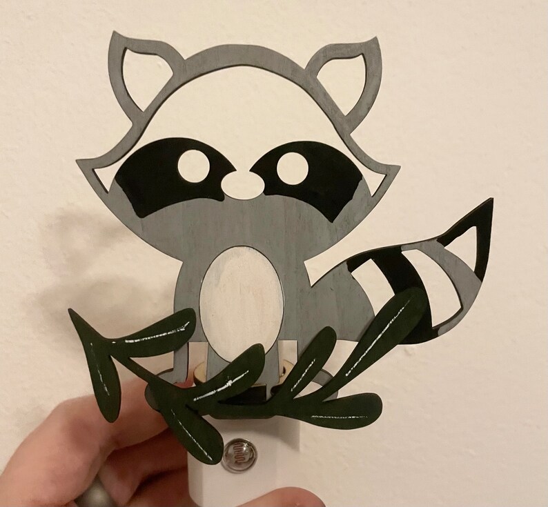 May include: A gray and black raccoon night light with a white belly and a green leaf design. The raccoon is holding a light bulb in its paws.