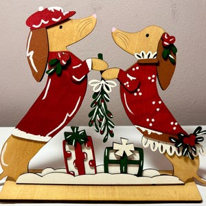 May include: Wooden Christmas decoration featuring two dachshunds in red outfits, holding mistletoe. The dogs are standing on a wooden base with two gift boxes. The outfits have white trim and accents. The dachshunds have brown ears and noses.