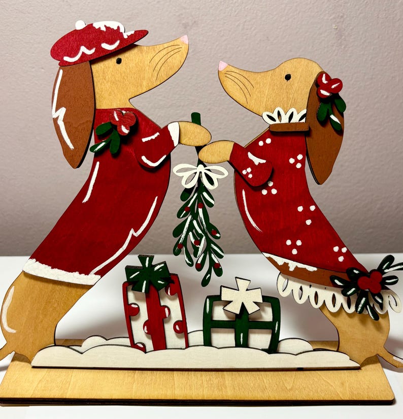 May include: Wooden Christmas decoration featuring two dachshunds in red outfits, holding mistletoe. The dogs are standing on a wooden base with two gift boxes. The outfits have white trim and accents. The dachshunds have brown ears and noses.