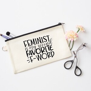 May include: A white canvas zippered pouch with a black zipper and black text that reads "Feminist is my second favorite f-word".