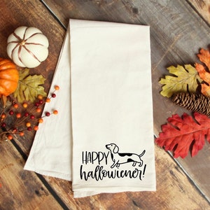 Pu&ograve; includere: Canovaccio bianco sporco con un'illustrazione di bassotto in bianco e nero e la scritta "Happy Hallowiener!". L'asciugamano &egrave; circondato da foglie autunnali, zucche e una pigna su una superficie di legno.