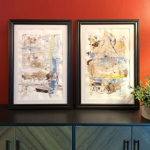 May include: Two framed abstract paintings with a white background and brown, blue, and yellow paint strokes. The paintings are in black frames and are hanging on a red wall.