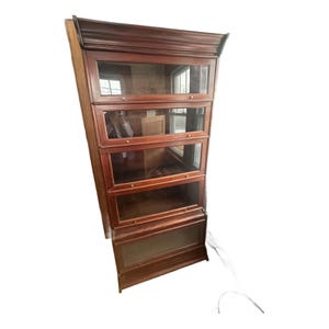 May include: A dark wood barrister bookcase with four glass-fronted sections. The bookcase has a traditional design with a crown molding top and brass hardware. The glass doors are framed in wood, and the bookcase is likely used for storing books or displaying items.
