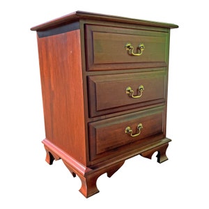 May include: A three-drawer nightstand made of dark wood with brass handles. The nightstand has a traditional design with slightly curved legs.
